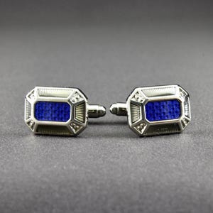May include: A pair of silver cufflinks with a rectangular blue center. The cufflinks have a geometric design with a textured border and a t-bar closure. The blue center has a textured pattern.
