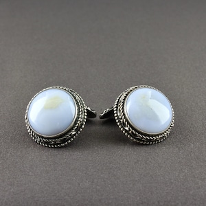 Op de afbeelding: Een paar zilveren manchetknopen met grote, ronde, lichtblauwe cabochon stenen. De stenen zijn gezet in een zilveren rand met een gedraaid touwontwerp. De manchetknopen hebben een klassiek, elegant ontwerp.