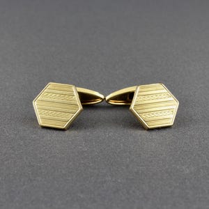 May include: A pair of gold-colored cufflinks. Each cufflink has a hexagonal face with a textured pattern of horizontal lines and a central zig-zag design. The cufflinks are set against a dark gray background.