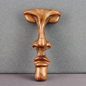 May include: A handcrafted wooden bottle stopper with a unique face design. The stopper features a detailed nose, mouth, and brow, all carved from a light brown wood. The stopper is approximately 5 inches tall.