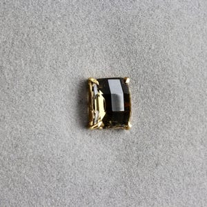 Peut inclure: Une pierre précieuse carrée et sombre avec un sertissage doré. La pierre précieuse a une forme rectangulaire avec une couleur sombre et fumée et une surface réfléchissante. Le sertissage doré a un design simple et élégant. Le bijou est posé sur un fond gris.
