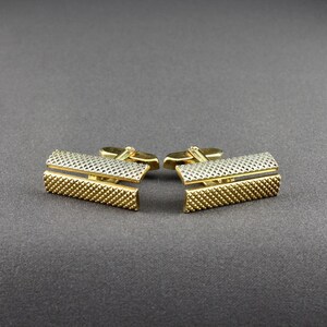 Könnte beinhalten: Ein Paar goldfarbene Manschettenknöpfe mit einem strukturierten, rechteckigen Design. Die Manschettenknöpfe haben eine goldfarbene Basis mit einem silberfarbenen oberen Abschnitt. Das strukturierte Muster ist ein sich wiederholendes Raster aus kleinen Quadraten. Die Manschettenknöpfe werden auf einer dunkelgrauen Oberfläche präsentiert.