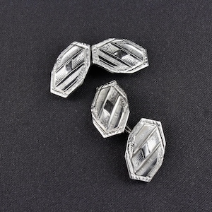 May include: Four silver cufflinks with an Art Deco design. Each cufflink has an elongated, geometric shape with etched lines and a central diamond shape. The cufflinks are displayed on a dark gray textured surface.