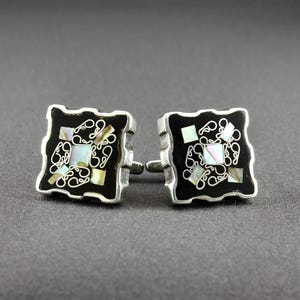 May include: A pair of square cufflinks with a black background and silver trim. The center of each cufflink features a decorative design with white and iridescent accents. The cufflinks are displayed on a gray surface.