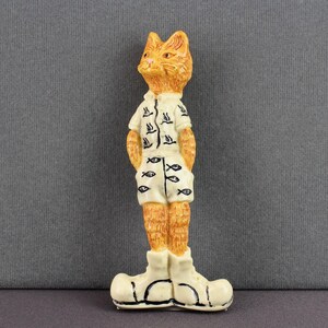May include: A figurine of a cat-like character wearing a white shirt and shorts with black fish and bird designs. The cat has orange fur and is wearing white boots. The figurine is standing with its hands in its pockets.