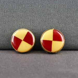 May include: Two round, gold-rimmed earrings. Each earring features a segmented design with alternating red and yellow sections. The earrings have a glossy finish and are set against a gray background.