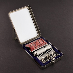 May include: A silver travel shaving kit with a red and silver patterned razor, a silver razor blade holder, and a silver comb. The kit has a built-in mirror.