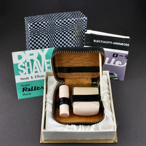May include: A black leather travel case with a zipper closure. The case contains a white plastic roller shaver and a small white plastic container. The case is open and the shaver and container are visible. The case is labeled "Dry Shave Handy & Efficient Pocket Roller Shaver".