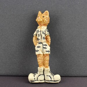 May include: A whimsical figurine of a cat wearing a short-sleeved shirt and shorts, both adorned with fish motifs. The cat is in cream-colored shoes. The figurine is tan and cream-colored, standing against a gray background.