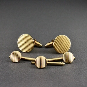 May include: A set of gold-toned metal cufflinks and matching shirt studs. Each piece features a circular face with a textured pattern of fine, parallel horizontal lines. The cufflinks have a hinged toggle mechanism, while the studs have a post.