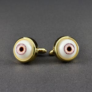 May include: A pair of gold-toned cufflinks, each featuring a detailed eye design. The eyes have a white background with a pink iris and a dark brown pupil. The cufflinks are round with a polished finish.