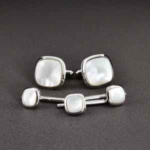 May include: A set of two silver-toned cufflinks and three matching shirt studs, each featuring a square-shaped, iridescent white mother-of-pearl inlay with rounded corners and a polished metallic frame, presented as a coordinated formal accessory set.