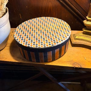 May include: A round, lidded box featuring a top surface decorated with a repeating pattern of small orange, black, and white polka dots. The sides of the box are adorned with vertical stripes in alternating shades of orange, black, and white.