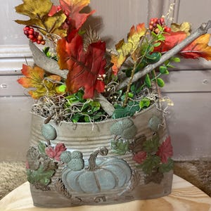 May include: A decorative metal container featuring a raised relief of a pumpkin, green leaves, and acorns. The container is filled with artificial autumn foliage, including red and yellow maple leaves, small red berries, and thin wooden branches.