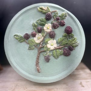 Puede incluir: Una pieza decorativa redonda de color verde azulado claro con un relieve en relieve de una rama de zarzamora. La rama tiene hojas verdes, flores blancas y moras de color púrpura oscuro. La pieza probablemente está hecha de un material duradero.