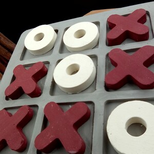 May include: A gray and white tic-tac-toe game board with red and white game pieces. The board has nine squares, and the pieces are arranged in a tic-tac-toe pattern.