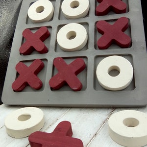 May include: A wooden tic-tac-toe game with red X's and white O's. The game board is gray and has nine squares. There are three red X's and three white O's on the board. There is one red X and one white O off the board.