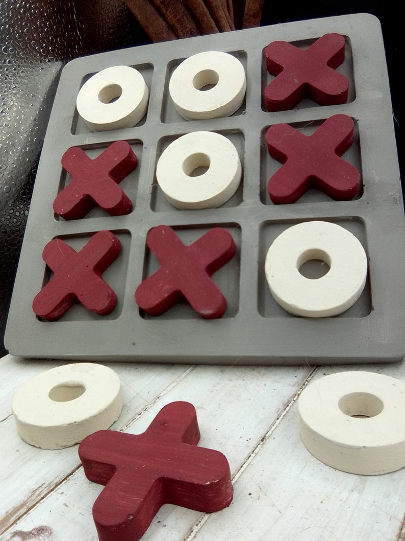 May include: A wooden tic-tac-toe game with red X's and white O's. The game board is gray and has nine squares. There are three red X's and three white O's on the board. There is one red X and one white O off the board.