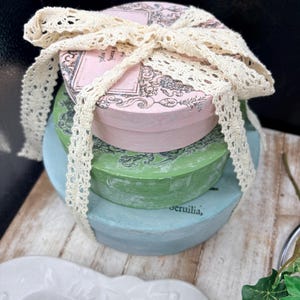 May include: A stack of three round decorative boxes in pastel colors: light blue, green, and pink. The top box is tied with a cream-colored lace ribbon. The boxes have a vintage aesthetic, with printed designs on the lids.