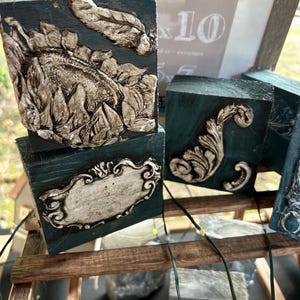 May include: Four teal blue wooden blocks with decorative white raised designs. The designs include a sunflower, a scroll, a frame, and a floral pattern. The blocks are arranged in a row on a wooden shelf.
