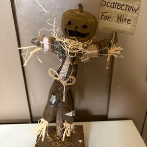 May include: A scarecrow figurine with a carved pumpkin head, wearing a brown plaid shirt and dark trousers. It features straw accents at the neck and wrists, stands on a square wooden base, and holds a sign that reads Scarecrow For Hire.