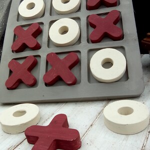 May include: A wooden tic-tac-toe game with red X's and white O's. The game board is gray and has nine squares. The pieces are arranged in a winning pattern for the X's. There are two extra pieces on the table, one red X and one white O.
