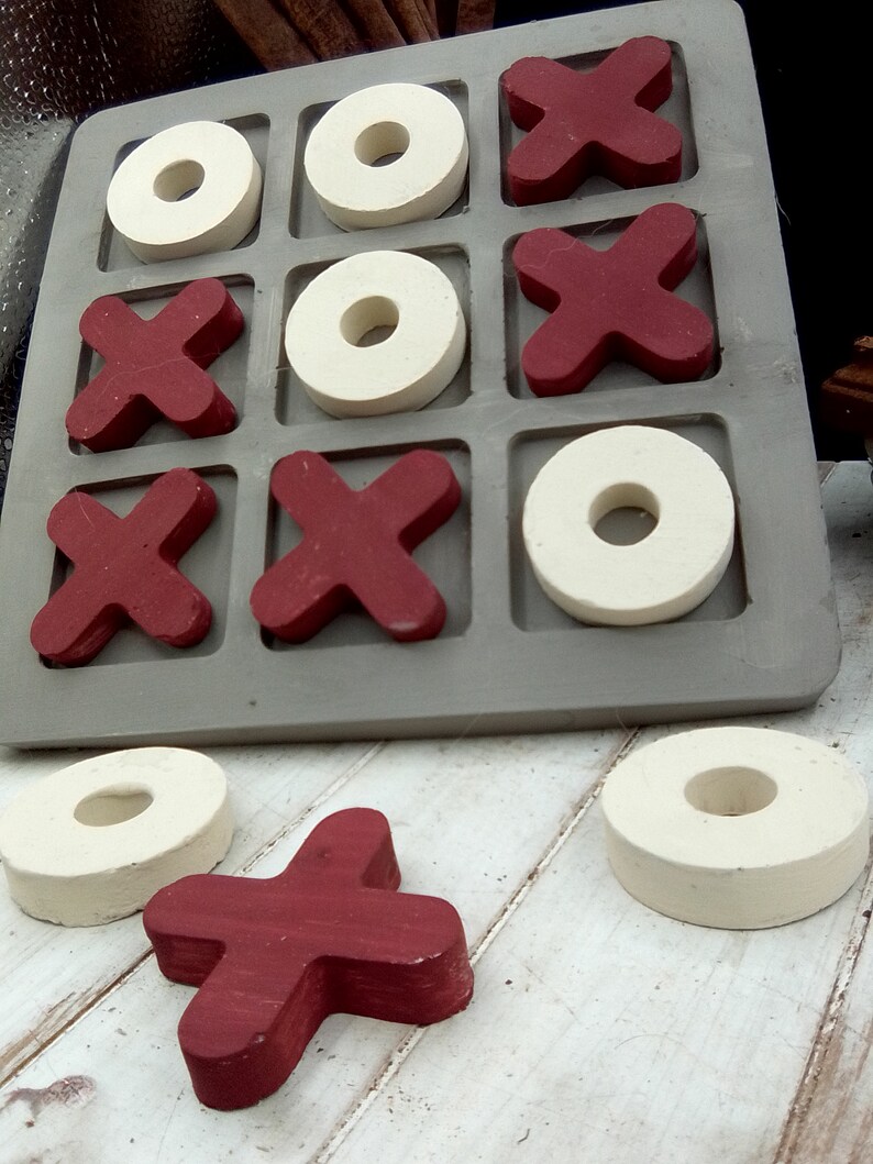 May include: A wooden tic-tac-toe game with red X's and white O's. The game board is gray and has nine squares. The pieces are arranged in a winning pattern for the X's. There are two extra pieces on the table, one red X and one white O.
