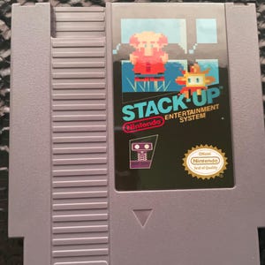 May include: A gray Nintendo Entertainment System game cartridge for the game Stack-Up. The label features pixel art of a character and a robot, with the title Stack-Up in teal text, the Nintendo logo, and an official Nintendo Seal of Quality.