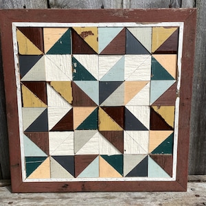 May include: A framed geometric wall art piece made of wood with a patchwork design. The wood is painted in shades of brown, yellow, green, white, and gray. The frame is a dark red color with a white inner border.