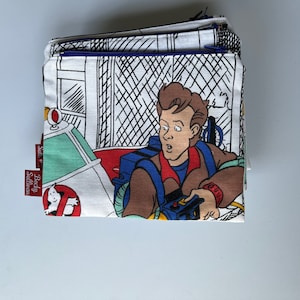 May include: A folded fabric item featuring a Ghostbusters-themed print. The design includes a cartoon image of a Ghostbuster, the Ecto-1 car, and the Ghostbusters logo. The item has a blue zipper and tags that read "Becky Sullivan".