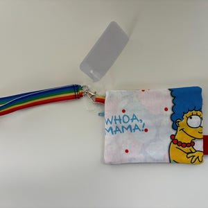 May include: A small, rectangular wallet featuring Marge Simpson with blue hair and a yellow complexion. The wallet has the phrase "WHOA, MAMA!" printed on it. A rainbow-striped wrist strap is attached to the wallet.