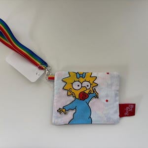 May include: A small, square, white fabric wallet featuring a cartoon image of Maggie Simpson in a blue dress. The wallet has a rainbow-striped wrist strap and a red tag that says "Becky Niff".