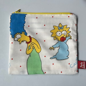 May include: A white zippered pouch with a yellow zipper pull. The pouch features a cartoon image of two yellow characters from the Simpsons, Marge and Maggie, on a white background with red polka dots. The text "CAR" is visible on the pouch. The pouch has a red label with the text "Becky Sullivan" on it.