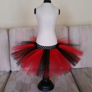 Può includere: Una gonna tutu di tulle rosso e nero con una cintura a pois neri e bianchi. La gonna è esposta su un manichino bianco.