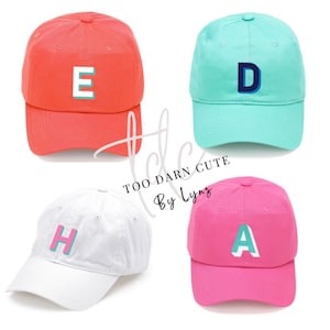 May include: Four baseball caps in different colors with a large letter embroidered on the front. The caps are red, white, pink, and turquoise. The letters are turquoise, pink, turquoise, and blue.