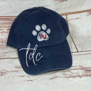 May include: A navy blue baseball cap with a white paw print embroidered on the front. The paw print has the initials "NW" in red inside.