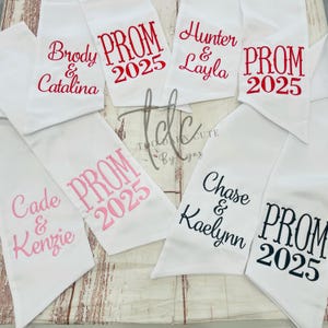 May include: White satin prom sashes embroidered with names and "PROM 2025" in various colors. The sashes are personalized with names like "Brody & Catalina," "Hunter & Layla," "Cade & Kenzie," and "Chase & Kaelynn."