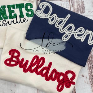 May include: Three t-shirts with team names. One white shirt has "Bulldogs" in red script. A navy blue shirt has "Dodgers" in white script. A white shirt has "Nets" in green glitter script.