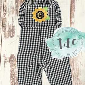 May include: Black and white gingham overall romper with a sunflower applique featuring the letter E. The romper has ruffles around the neckline and the legs. The romper is for a baby or toddler.