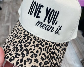 May include: A white baseball cap with the embroidered phrase "LOVE YOU, mean it." The brim features a tan and black leopard print. The text "TOO DARN CUTE BY LYNZ" is printed at the bottom.