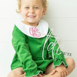 May include: A little girl wearing a green dress with a white Peter Pan collar and pink monogram. The dress has white lace trim on the sleeves and collar. The girl is wearing a pink bow in her hair. The text "Too Darn Cute By Lynnz" is visible on the dress.
