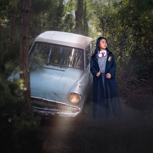May include: A young person wearing a dark blue robe with a red and gold tie, holding a wand, stands in front of a vintage blue car in a wooded area.