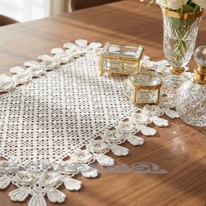 Puede incluir: Camino de mesa blanco, tejido a ganchillo, con diseño floral y bordes festoneados. Se encuentra sobre una mesa de madera, junto a un jarrón de cristal con rosas blancas y cajas de cristal decorativas.