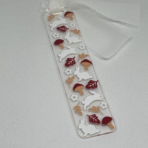 May include: A white acrylic bookmark with a white ribbon. The bookmark features a repeating pattern of white bunnies, red mushrooms, and brown leaves.