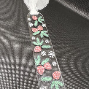 May include: A clear plastic bookmark with a winter theme. The bookmark features red and green pine cones, green pine branches, and white snowflakes.