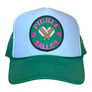 May include: A green and white trucker hat with a circular patch. The patch features the words "PICKLE BALLER" in pink, along with pickleball paddles and a ball. The hat has a green brim and mesh back.
