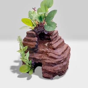 May include: A brown, terraced rock formation with a small cave opening, adorned with green and purple artificial plants. The rock structure has a textured surface and is designed to resemble a natural habitat.