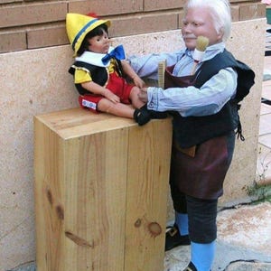 Può includere: Una bambola di Pinocchio è seduta su un blocco di legno mentre una persona vestita da Geppetto le spazzola il piede. La bambola di Pinocchio indossa un cappello giallo, un gilet nero e pantaloncini rossi. La figura di Geppetto indossa una camicia blu, un gilet nero e pantaloni marroni.