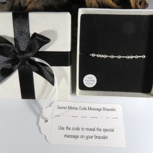 May include: A cream-coloured gift box with a black ribbon, alongside an open box displaying a silver Morse code bracelet on a black background. A tag reads "Secret Morse Code Message Bracelet" and "Use the code to reveal the special message."