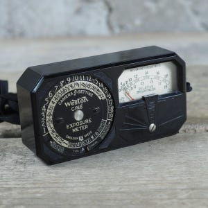 Puede incluir: Un fotómetro Weston Cine negro con marcas blancas y rojas. Cuenta con un dial circular con números y ajustes, y una ventana rectangular que muestra el brillo relativo. Una correa negra está adjunta.